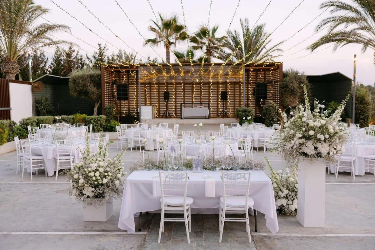 Evening wedding reception at Alassos with a fairy-light canopy strung above white-linen round tables, white chiavari chairs, tall white ground florals and palm trees against a dusk sky by the sea