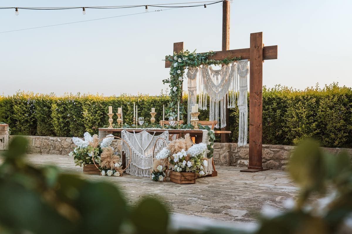 Boho sweetheart table at Liopetro with a macramé backdrop hung from a timber frame, trailing eucalyptus and white roses, pampas grass in wooden crates and gold candlesticks, against a stone wall at dusk
