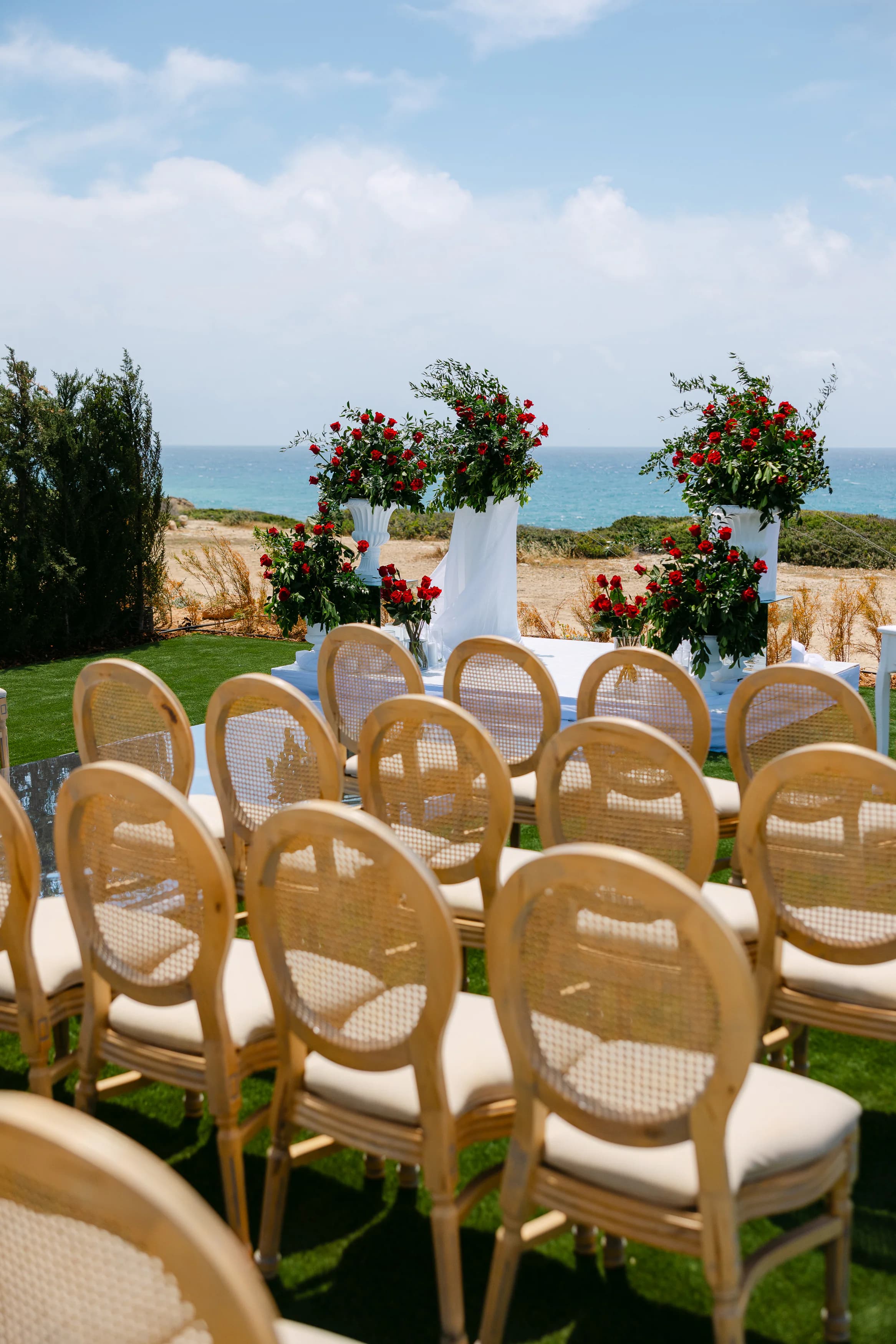 Wedding ceremony altar at OZEA with rows of cane Louis-back chairs facing a white draped table and tall red rose and foliage arrangements raised on white plinths, with the open sea behind