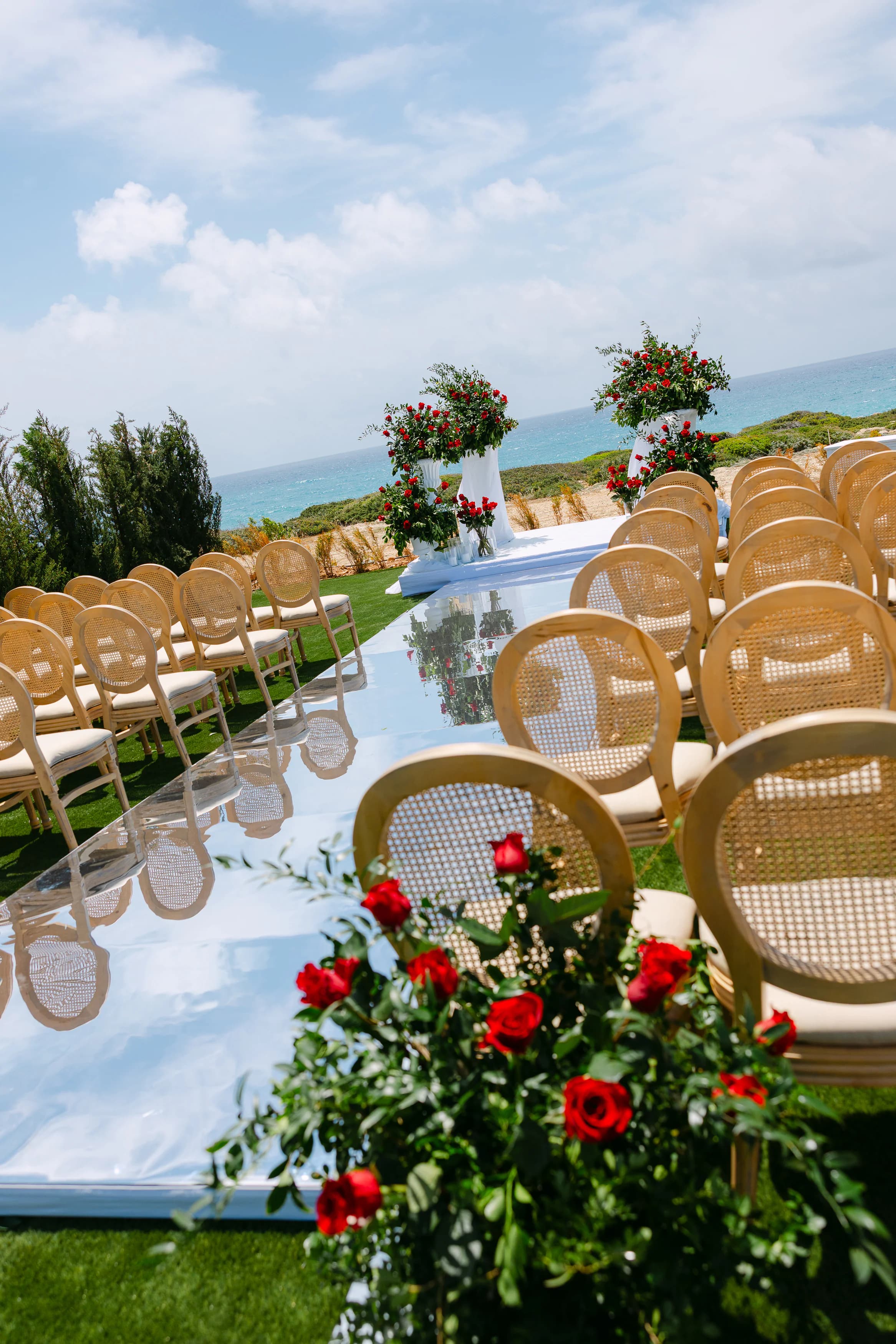 Mirrored ceremony aisle at OZEA framed by cane chairs and lush red rose arrangements with sea views beyond