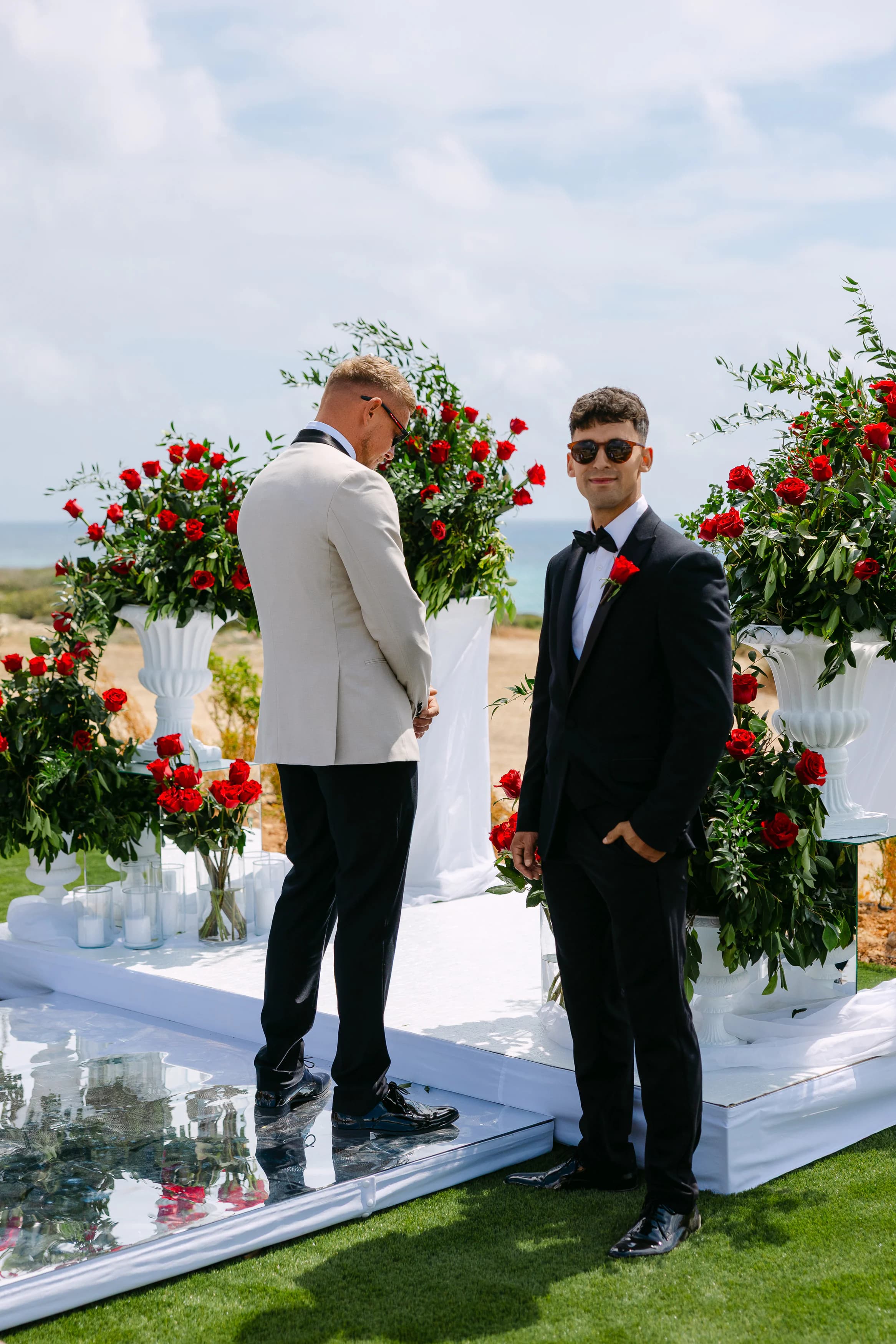 Groom and best man waiting at the OZEA ceremony altar surrounded by tall red rose arrangements with the Mediterranean Sea behind