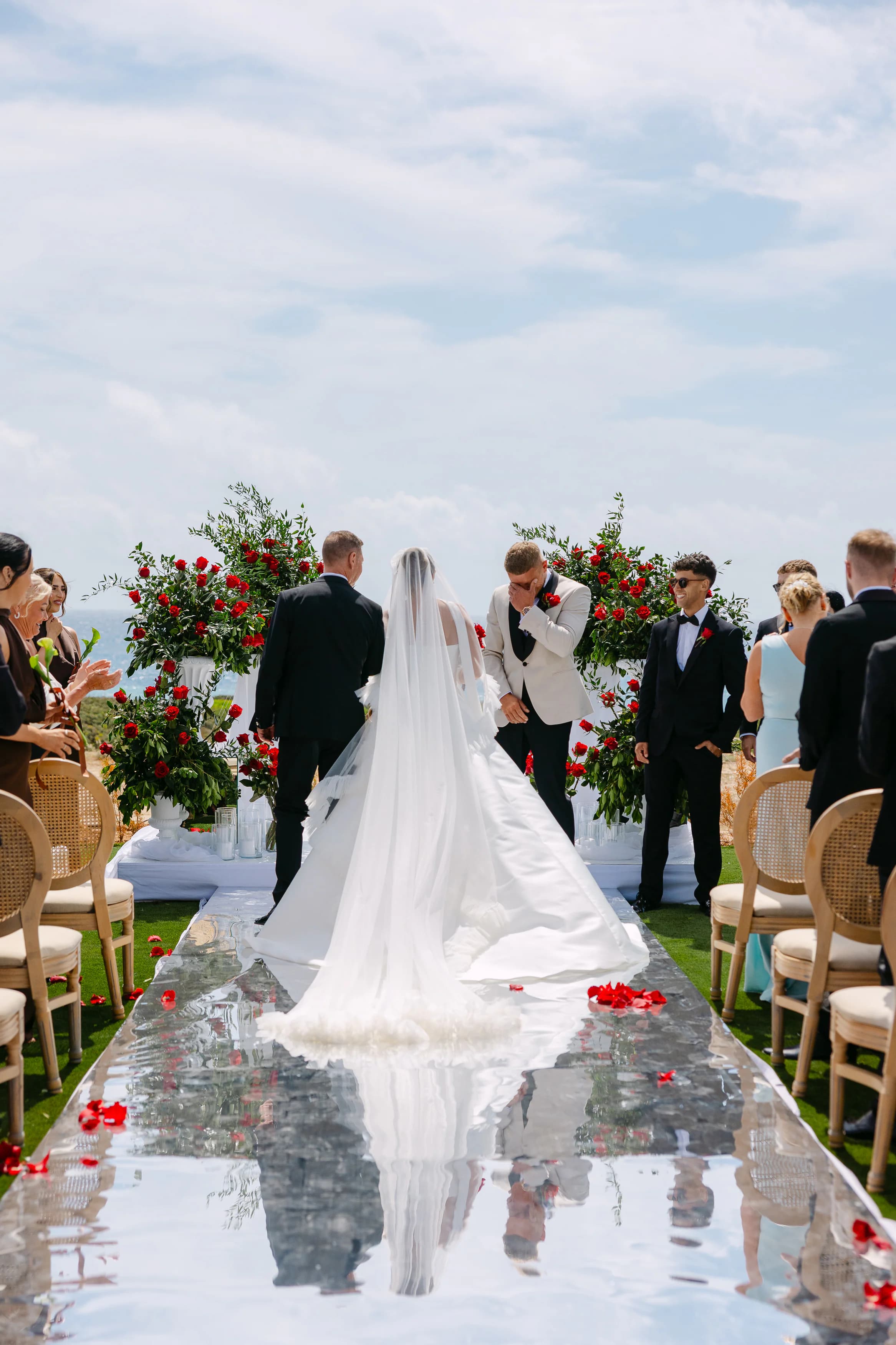 Bride walking down the mirrored aisle at OZEA with a long cathedral veil and rose petals scattered on the runner, Sea Caves Paphos