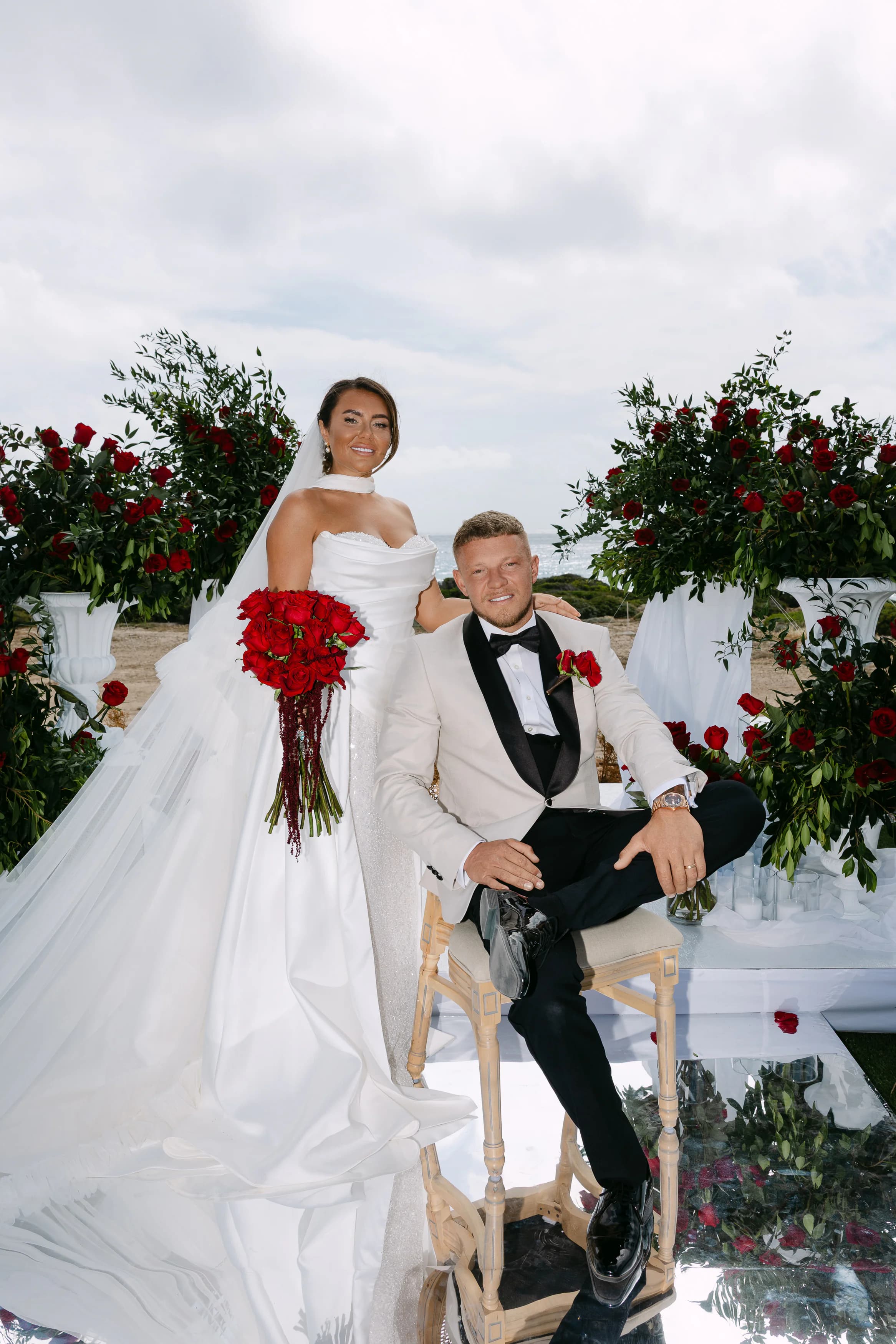 Bride and groom portrait at the OZEA ceremony space framed by red roses, classic black tuxedo and ivory ballgown, Paphos wedding venue