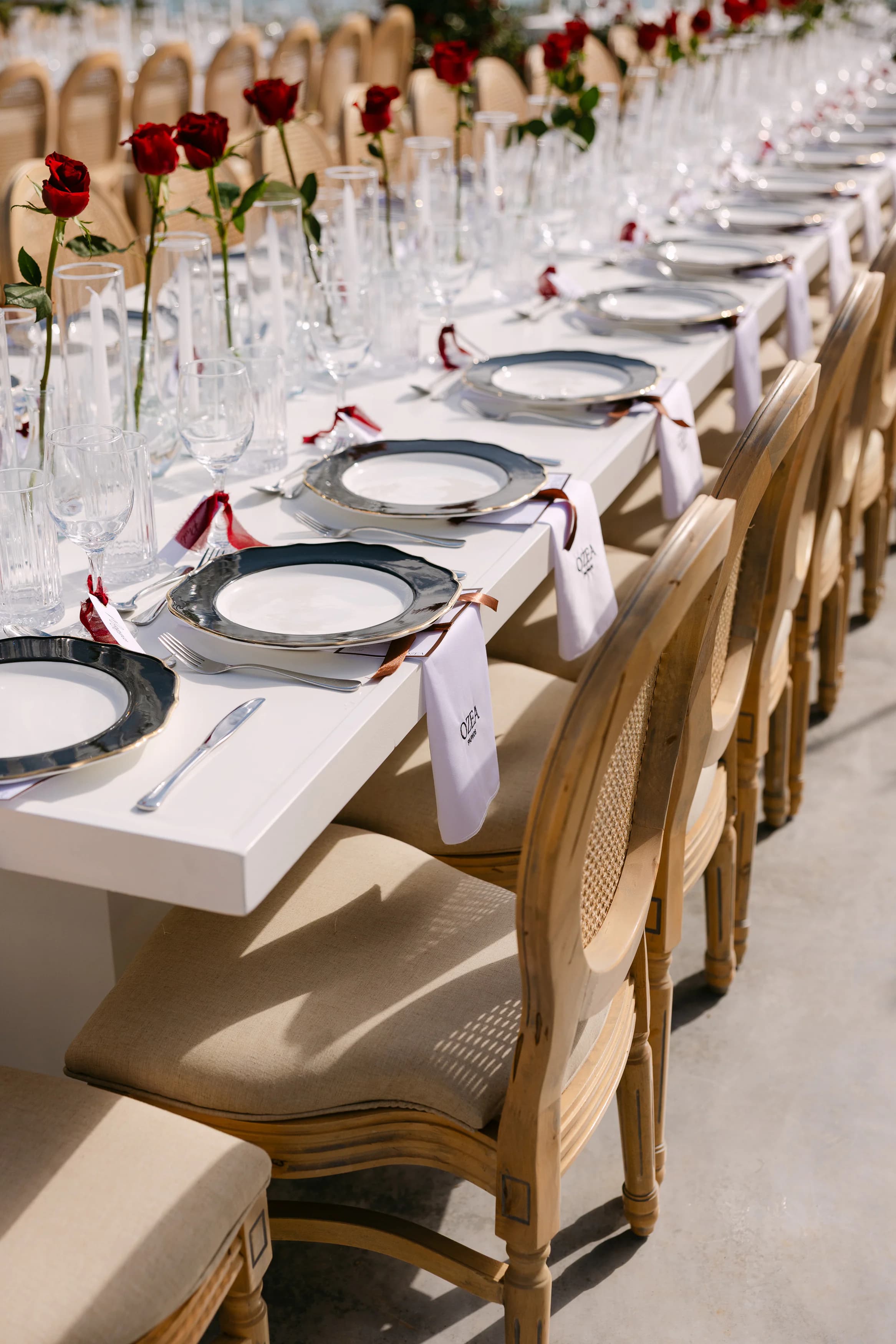 Close-up of wedding banquet table place settings at OZEA with silver-rimmed charger plates, gold-edged china, ribbon-tied embroidered napkins, monogrammed name cards, crystal stemware and single red roses in bud vases