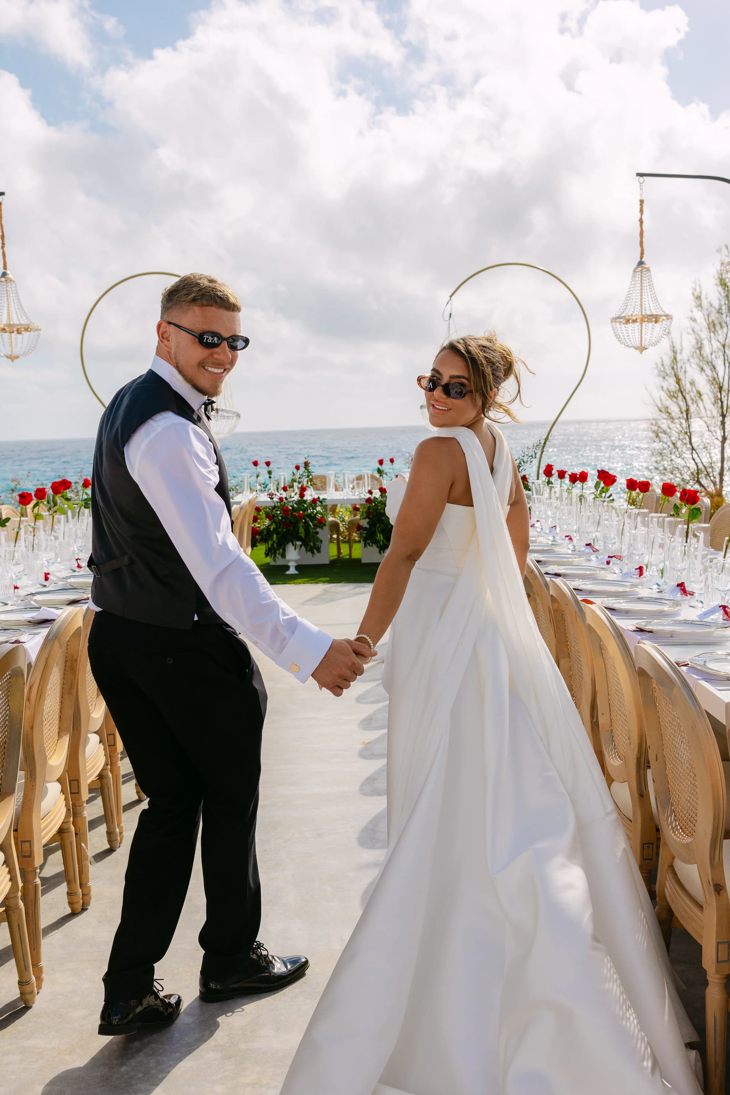 Newlyweds hand in hand walking toward the OZEA reception table with crystal chandeliers and the Mediterranean Sea behind them