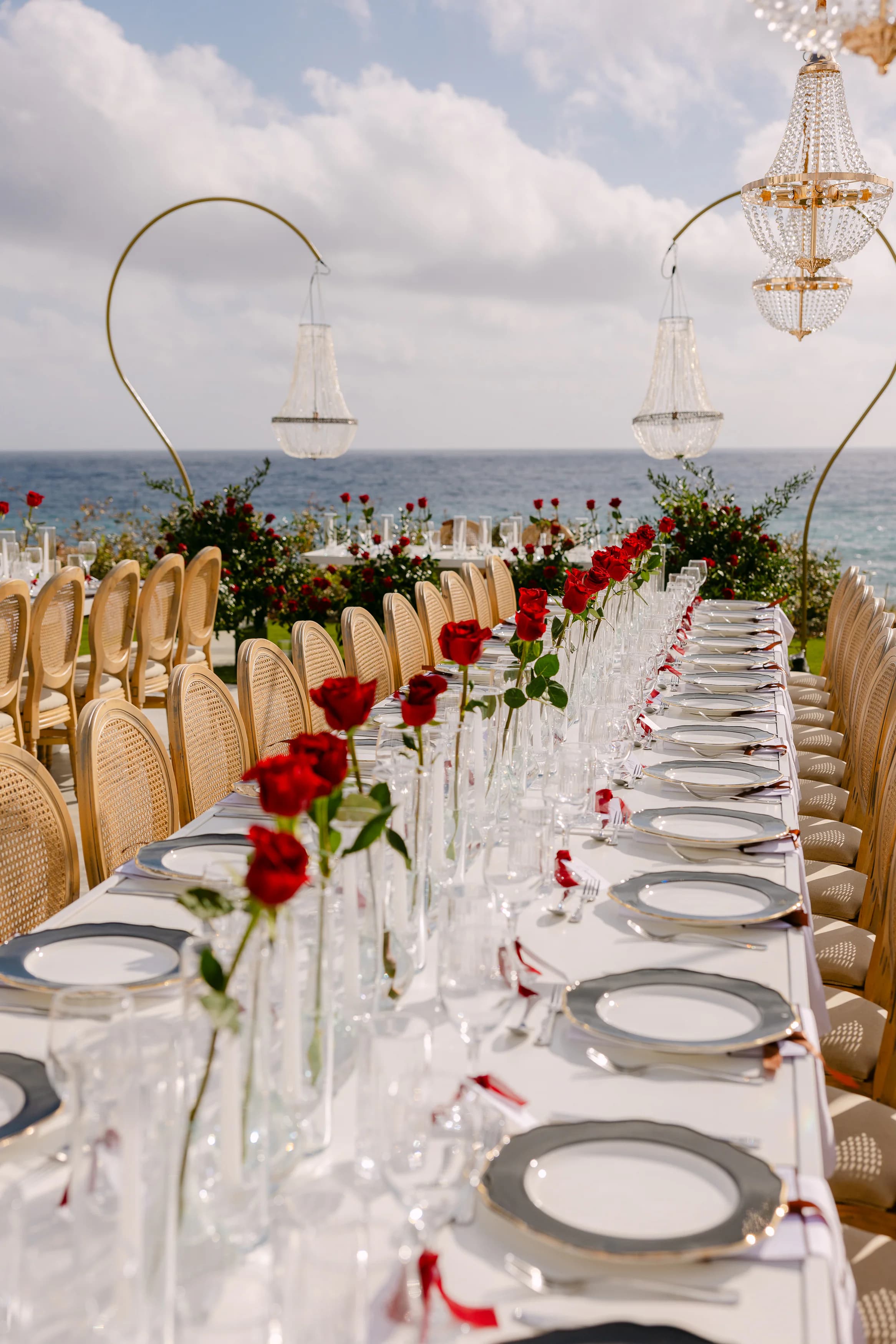 OZEA wedding banquet table styled with single-stem red roses, crystal stemware and silver-rimmed place settings against the sea