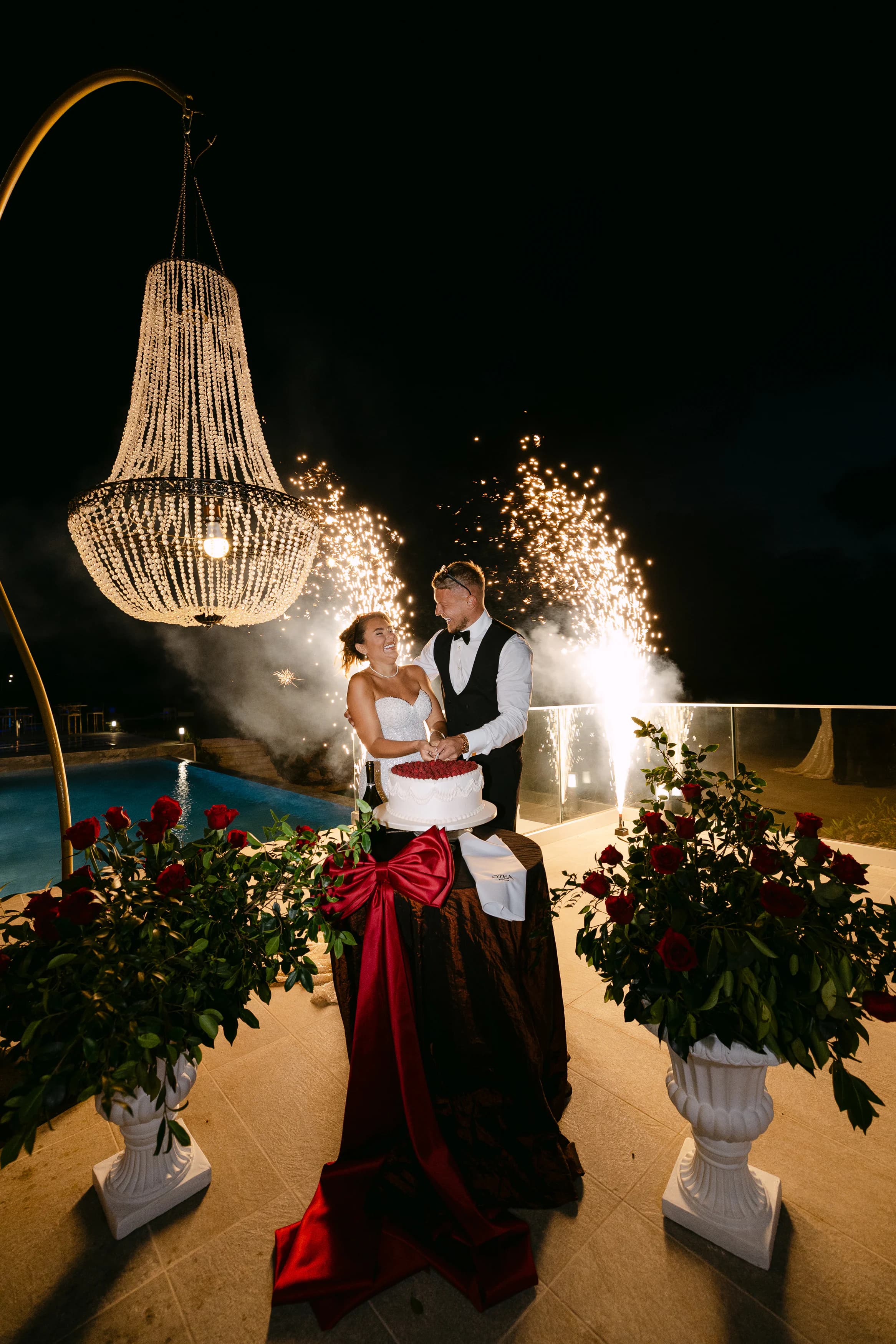 Bride and groom cutting the wedding cake at OZEA under sparkler fountains, with crystal chandelier and red rose urns at night